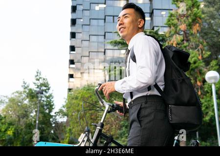 Vue latérale d'un jeune homme de bureau de rêve avec sac à dos et vélo, à l'écart des bâtiments urbains et des arbres Banque D'Images