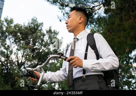 Vue latérale d'un jeune homme de bureau de rêve avec sac à dos et vélo, à l'écart des bâtiments urbains et des arbres Banque D'Images