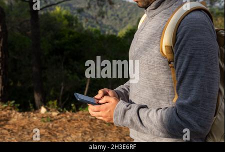 Vue latérale de jeune homme anonyme dans des vêtements décontractés et des messages sac à dos sur smartphone debout dans une forêt verdoyante pendant le trekking Banque D'Images