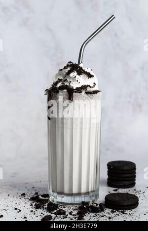 Verre de milkshake blanc sucré recouvert de crème fouettée et de chapelure de biscuits au chocolat servi avec de la paille sur une table désordonnée près du mur Banque D'Images