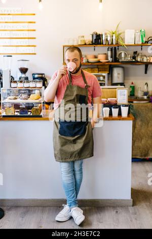 Tout le corps d'un barista masculin sérieux en tablier regardant l'appareil photo tout en buvant du café chaud près du comptoir dans une maison de café moderne Banque D'Images