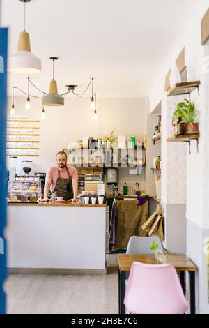 Un barista masculin en tablier souriant et regardant l'appareil photo tout en se tenant au comptoir avec une tasse de café dans le café Banque D'Images