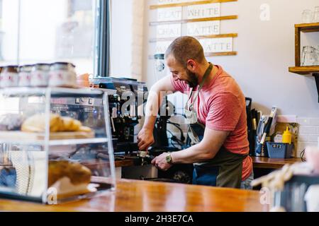 Vue latérale du barista masculin en tablier avec porte-filtre préparant du café à l'aide d'une machine à café tout en se tenant près du comptoir dans la maison à café Banque D'Images