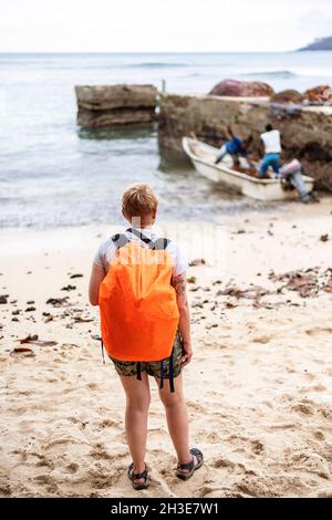 Vue arrière du voyageur anonyme en vêtements décontractés et sac à dos orange debout sur la plage de sable et regardant les pêcheurs locaux en bateau Banque D'Images