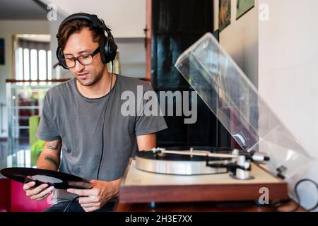 Homme concentré dans des lunettes écoutant de la musique dans les écouteurs du lecteur dans l'appartement spacieux Banque D'Images