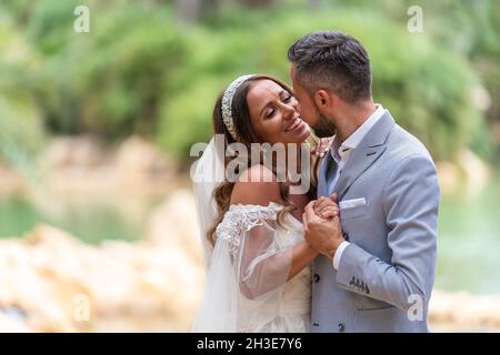 Vue latérale de jeunes mariés heureux dans des vêtements de mariage debout et tenant les mains tandis qu'embrassé près du lac et des arbres verts dans la journée dans le jardin Banque D'Images