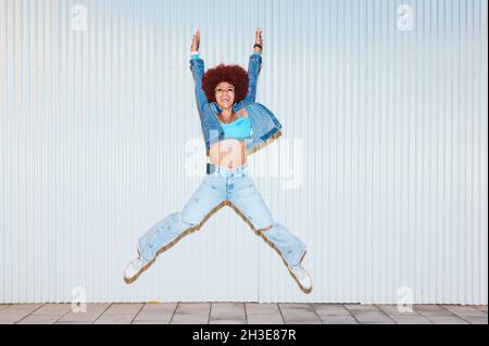 Corps complet de femme gaie avec une coiffure afro portant une tenue élégante sautant avec des jambes et des bras surélevés sur fond blanc dans la rue Banque D'Images