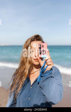 Jeune femme en vêtements mouillés prenant des photos sur l'appareil photo tout en regardant l'appareil photo sur une plage de sable près de la mer agitant Banque D'Images