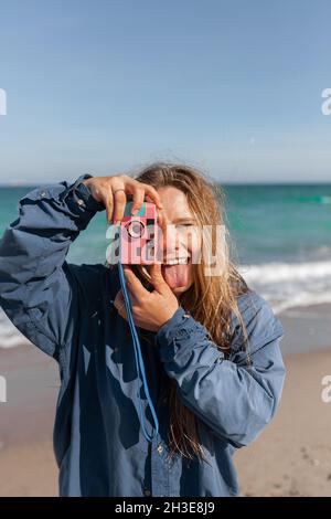 Jeune femme gaie en vêtements mouillés prenant des photos sur l'appareil photo tout en se tenant debout avec la langue sur la plage de sable près de la mer agitant Banque D'Images