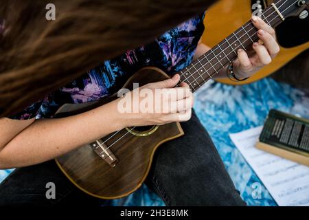 Petite musicienne féminine de talent non reconnaissable avec cheveux bruns dans des vêtements décontractés jouant ukulele et chantant la chanson tout en étant assis sur une plage de sable à nat Banque D'Images