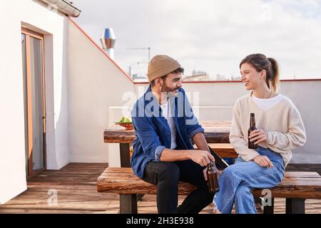 Jeunes copains positifs, hommes et femmes, assis au banc avec des bouteilles de bière et ayant une conversation agréable sur la terrasse Banque D'Images