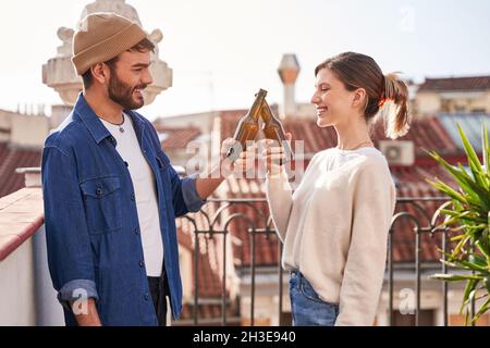 Des jeunes amis enchantés dans des vêtements décontractés qui se clinquent des bouteilles de bière tout en se rafraîchit sur la terrasse ensemble Banque D'Images