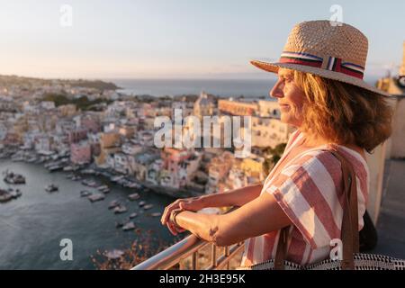 Vue latérale d'une touriste féminine paisible dans une robe élégante et chapeau de paille en admirant la vue pittoresque de l'île de Procida sous le ciel du coucher du soleil Banque D'Images