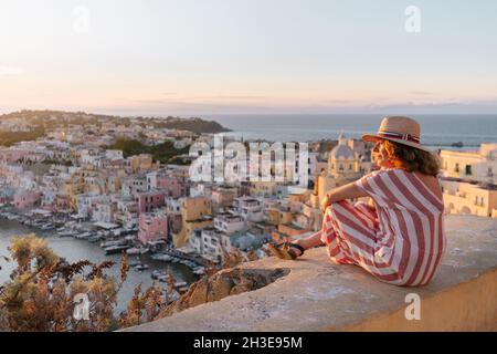 Vue latérale d'une touriste féminine paisible dans une robe élégante et chapeau de paille assis sur la frontière de pierre au sommet de la colline et en admirant la vue pittoresque de l'île de Procida Banque D'Images