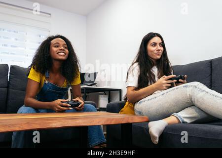 Des amis féminins multiraciaux positifs assis sur un canapé et jouant à un jeu vidéo tout en passant du temps ensemble à la maison Banque D'Images