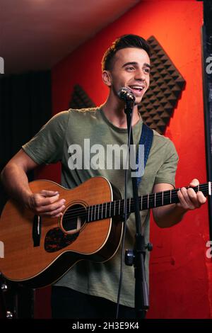 Positif jeune homme guitariste jouant de la guitare acoustique et chantant dans le microphone en club de lumière Banque D'Images