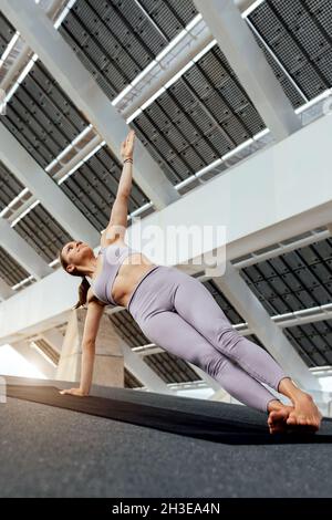 Femme pieds nus dans les vêtements de sport pratiquant Utthita Vasishthasana posture sur le tapis près du panneau photovoltaïque dans la rue pendant l'entraînement intense de yoga Banque D'Images