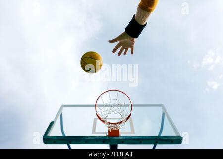 De dessous de crop anonyme personne jetant le ballon de basket-ball dans le cerceau tout en jouant sur le terrain de sport public contre le ciel bleu Banque D'Images