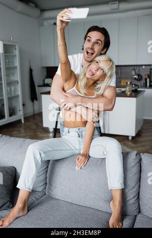 femme blonde qui colle à la langue tout en prenant le selfie avec un ami heureux dans le salon Banque D'Images