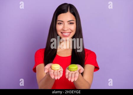 Photo de charmante jeune femme douce porter une tenue rouge souriante tenant deux macarons isolé couleur violet fond Banque D'Images