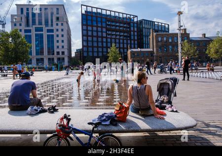 Les parents se détendent sur les bancs tandis que les enfants jouent dans l'eau aux fontaines de Granary Square, King's Cross, Londres. Banque D'Images