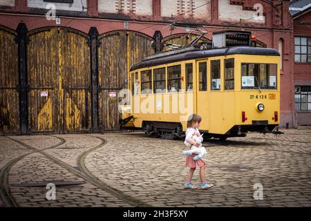 Wroclaw, Pologne - 19 septembre 2021: Une petite fille avec un jouet licorne, devant un tram jaune vintage. Bâtiment historique de dépôt de briques en arrière-plan. Banque D'Images