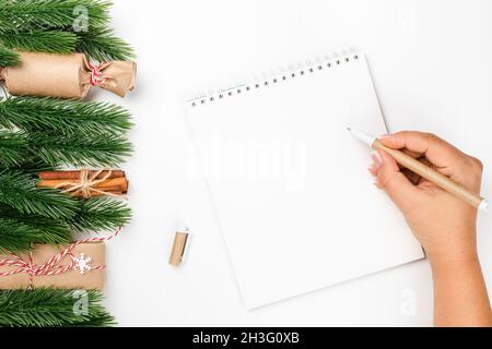 Femme écriture à la main dans bloc-notes blanc avec espace de copie avec stylo de travail sur table blanche avec des décorations faites de branches d'arbre de Noël et de cadeau Banque D'Images