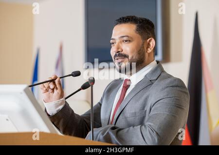Content politicien du Moyen-Orient confiant en costume gris debout sur le podium de la tribune et parlant en micro tout en présentant un rapport à la conférence Banque D'Images