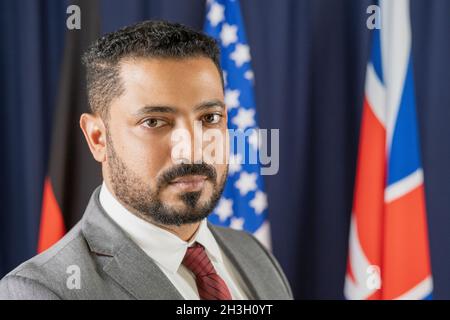 Portrait d'un homme politique barbu de race mixte sérieux en costume posant contre des drapeaux nationaux dans le hall des congrès Banque D'Images