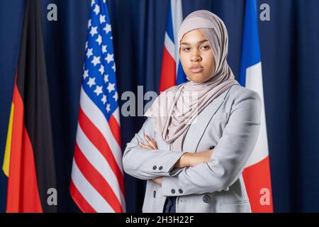 Portrait d’une femme musulmane sérieuse et confiante en veste officielle et hijab debout avec des armes croisées contre des drapeaux nationaux Banque D'Images