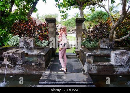 Portrait de la jeune femme musulmane asiatique portant le hijab pose dans le jardin avec étang et fontaine d'eau à Bali.Sourire et expression heureuse. Banque D'Images