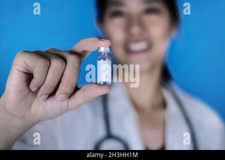Un médecin attrayant, portant un manteau blanc avec un stéthoscope autour de son épaule, tenant un flacon en verre de flacon de vaccin avec une étiquette blanche vierge, smili Banque D'Images