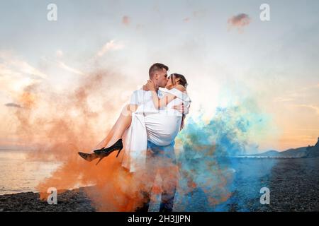 Mariage.Marié tient la mariée dans ses bras et l'embrasse, entouré de fumée colorée.Il y a un océan et un coucher de soleil en arrière-plan.Saint-Valentin Banque D'Images