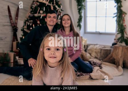 Portrait de famille aimante près de l'arbre de Noël avec des cadeaux sous lui.Fille avec maman et papa dans une chambre confortable décorée de vacances. Banque D'Images