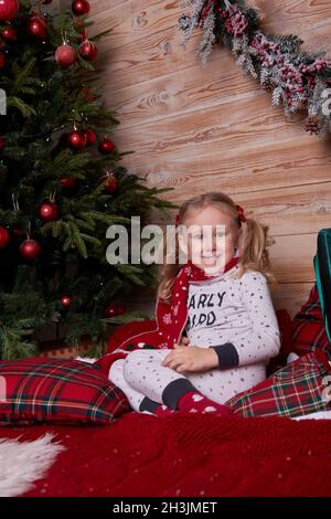 Portrait d'une fille en pyjama assise sur le lit sous un arbre de Noël décoré avec des cadeaux dans des boîtes-cadeaux.Vacances du nouvel an. Banque D'Images