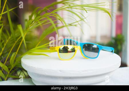 Lunettes de soleil jaunes et bleues sur terrasse blanche digue sur fond rose flou de maison Banque D'Images