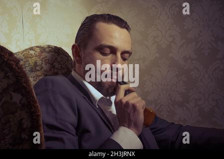 Jeune homme beau assis dans une chaise et fumant pipe sur fond vintage Banque D'Images