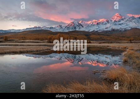 Incroyable lever de soleil d'automne avec des montagnes enneigées, forêt, mélèze doré, herbe couverte de givre, la première glace sur le lac et magnifique reflet Banque D'Images