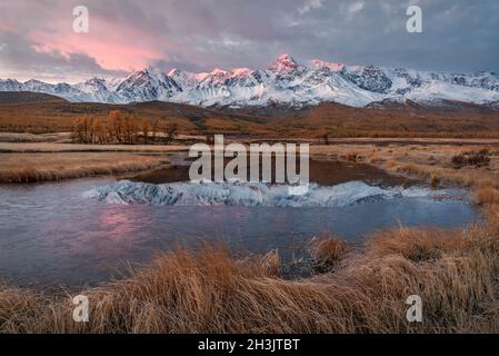 Incroyable lever de soleil d'automne avec des montagnes enneigées, forêt, mélèze doré, herbe couverte de givre, la première glace sur le lac et magnifique reflet Banque D'Images