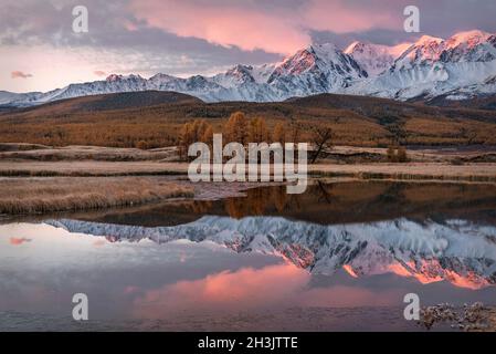 Incroyable lever de soleil d'automne avec des montagnes enneigées, forêt, mélèze doré, herbe couverte de givre, la première glace sur le lac et magnifique reflet Banque D'Images