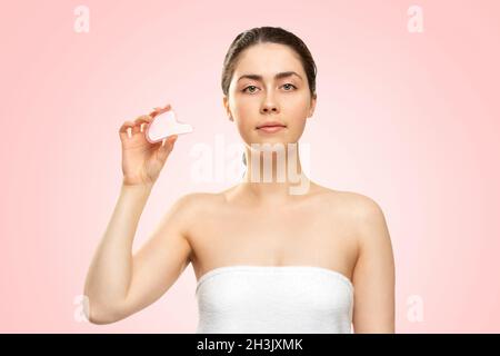 Massage Gua Sha.Une femme dans une serviette de bain blanche montre un grattoir de guasha de quartz.Fond rose.Copier l'espace.Concept de médecine alternative et de cosmétique Banque D'Images