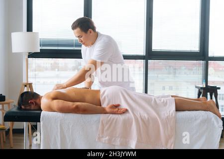 Photo de côté d'un masseur professionnel masculin avec de fortes mains tatouées massant le dos et l'épaule d'un sportif musclé allongé sur l'estomac. Banque D'Images