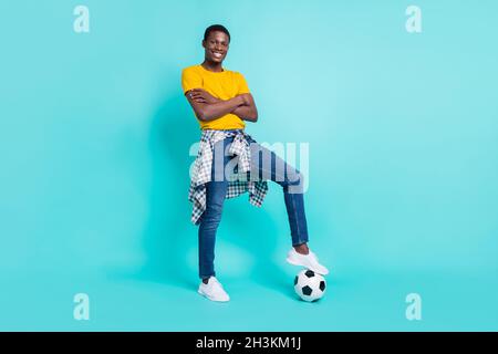 Photo pleine grandeur d'optimiste brunette hairdo jeune homme jouer football porter jaune t-shirt jeans baskets isolées sur fond de couleur cyan Banque D'Images
