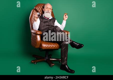 Photo de riche anglais gentleman assis chaise boisson fumée whisky cigare porter costume rayé sur fond vert Banque D'Images