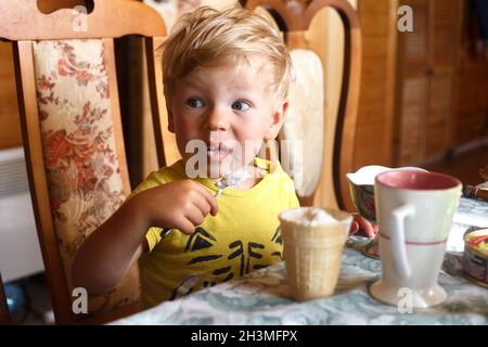 Un garçon blond s'assoit à la table et mange lui-même de la glace avec une cuillère Banque D'Images