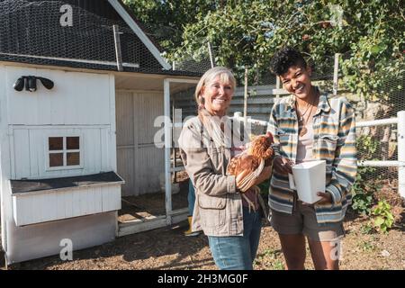 Portrait de la mère et de la fille multiraciales souriantes avec du poulet à la ferme le jour ensoleillé Banque D'Images