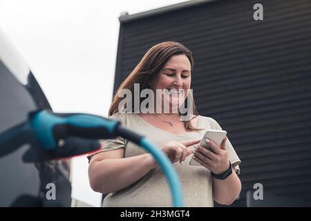 Femme mûre souriante utilisant un smartphone à la station de charge Banque D'Images