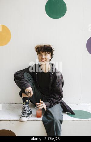 Portrait d'un adolescent souriant assis avec un verre sur le mur de soutènement Banque D'Images