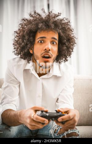 Les joueurs jouent à un jeu informatique à la maison, en isolement personnel.Surprise arabe jeune gay tient Play Station joystick assis au canapé.Comp Banque D'Images