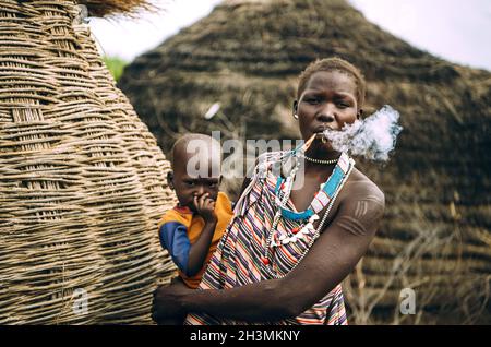 Tribu TOPOSA, SOUDAN DU SUD - 12 MARS 2020: Femme de la tribu Toposa fume et porte bébé tout en se tenant près de la tradition Banque D'Images